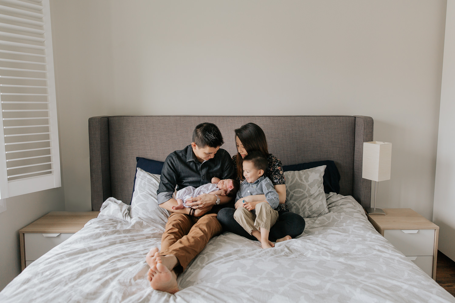 family of 4 sitting on master bed, dad holding 2 week old baby girl and mom with 3 year old toddler boy in her lap, everyone looking at new baby - Stouffville Lifestyle Photos