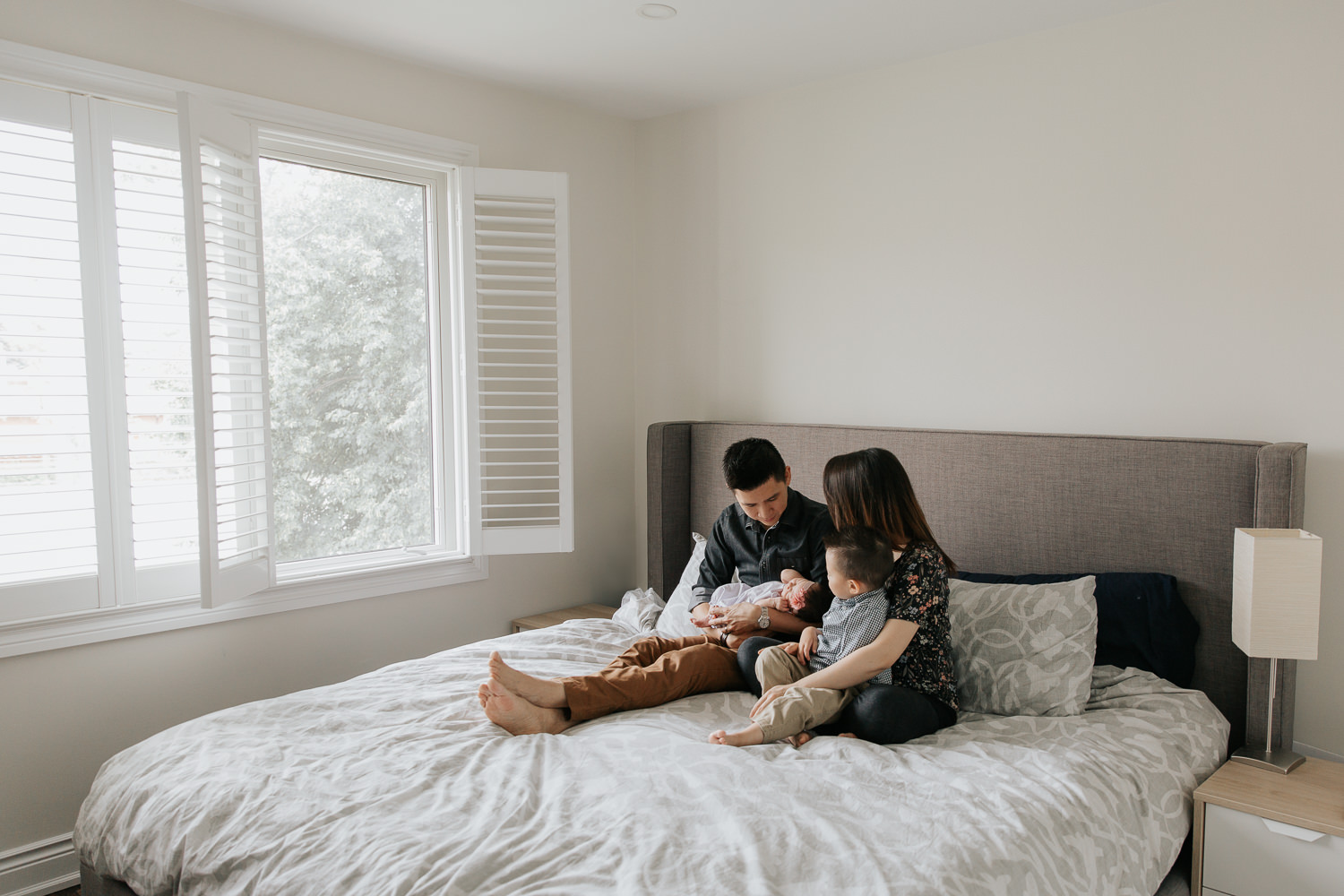 family of 4 sitting on master bed, dad holding 2 week old baby girl and mom with 3 year old toddler boy in her lap, everyone looking at new baby - Markham Lifestyle Photos