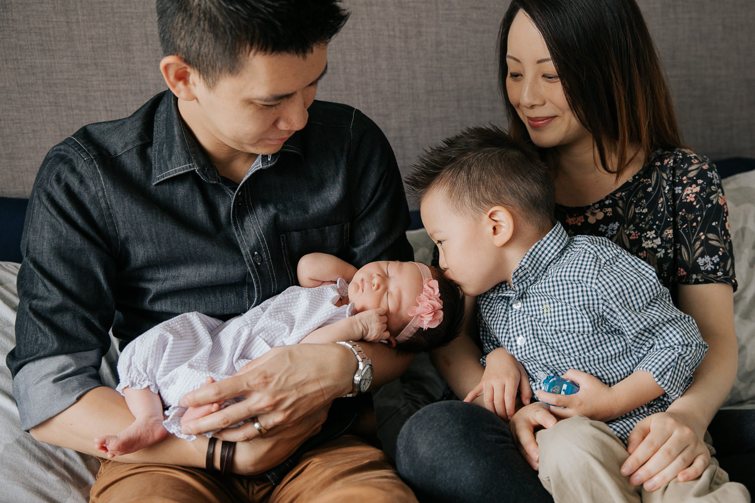 family of 4 sitting on master bed, dad holding 2 week old baby girl and mom with 3 year old toddler boy in her lap, big brother kissing little sister on head - Barrie Lifestyle Photos