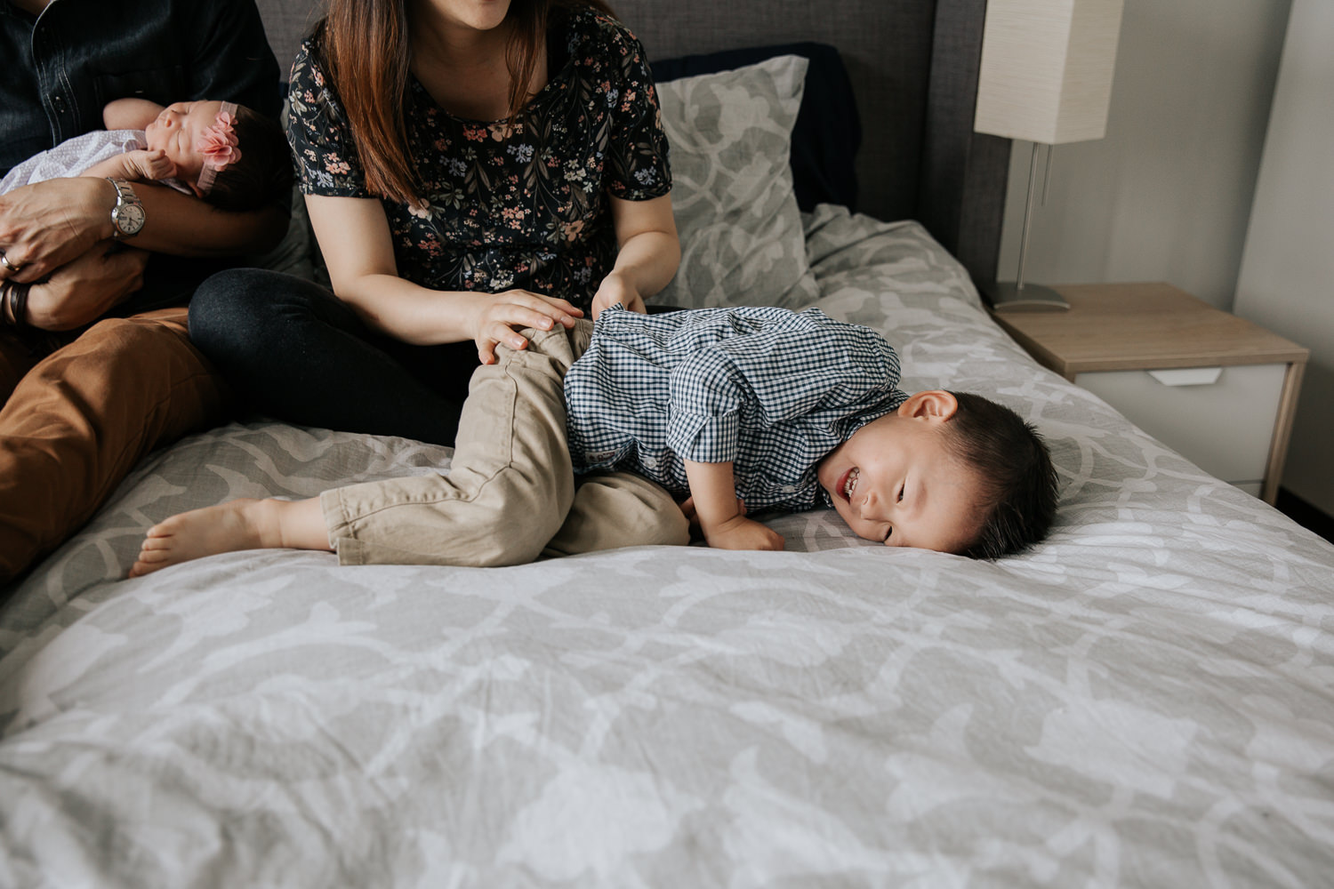 3 year old toddler boy lying on parent's bed laughing while mom tickles him, dad holding 2 week old baby sister in background - Newmarket Lifestyle Photography