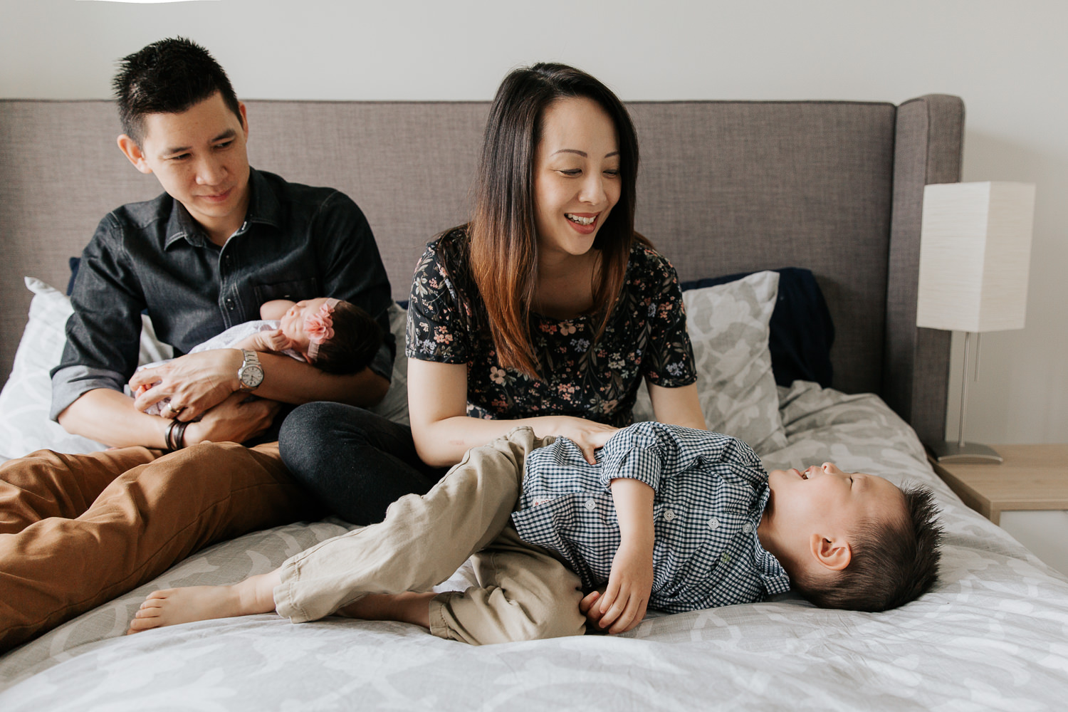 3 year old toddler boy lying on parent's bed laughing while mom tickles him, dad holding 2 week old baby sister in background - Stouffville Lifestyle Photography