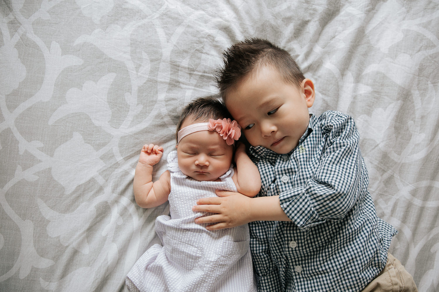 3 year old toddler boy lying on bed next to 2 week old sleeping baby sister, his hand on her torso - Markham In-Home Photos