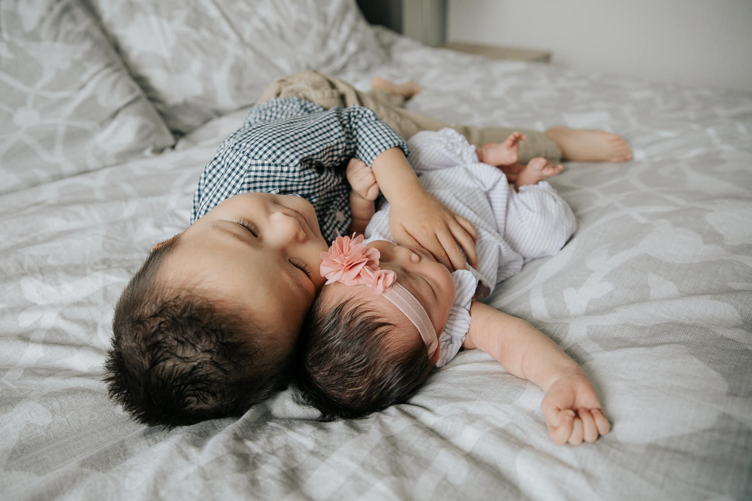 3 year old toddler boy lying on bed next to 2 week old sleeping baby sister, his hand on her torso - GTA In-Home Photos