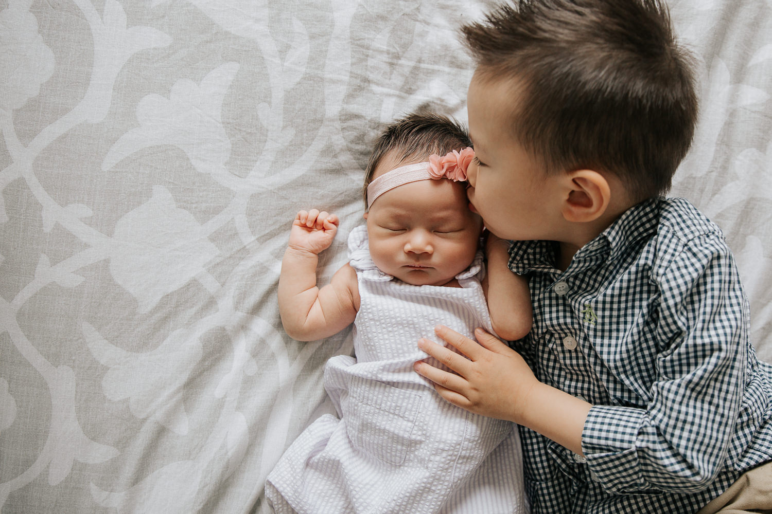 3 year old toddler boy lying on bed next to 2 week old sleeping baby sister, kissing her on cheek - Barrie In-Home Photos