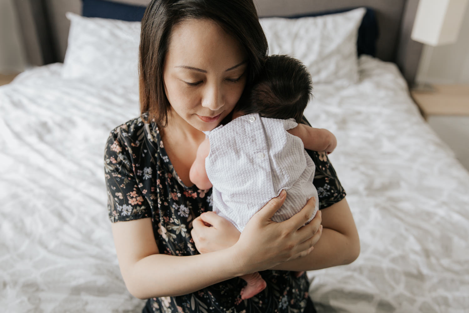 mom sitting on edge of bed snuggling sleeping 2 week old baby girl in white and purple striped outfit to her chest - Newmarket Lifestyle Photos