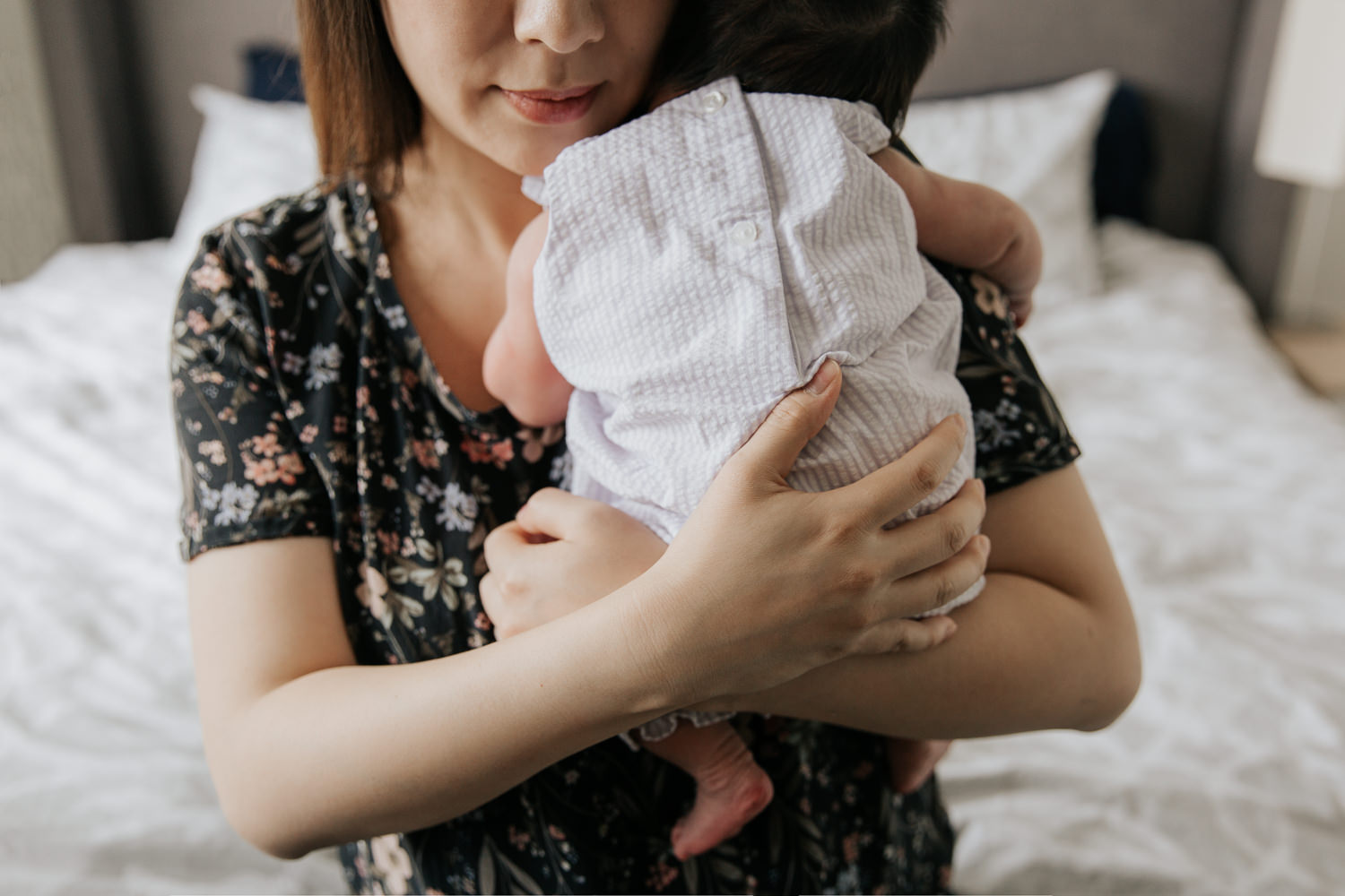 mom sitting on edge of bed snuggling sleeping 2 week old baby girl in white and purple striped outfit to her chest, close up of mother's hands holding daughter - Stouffville Lifestyle Photos