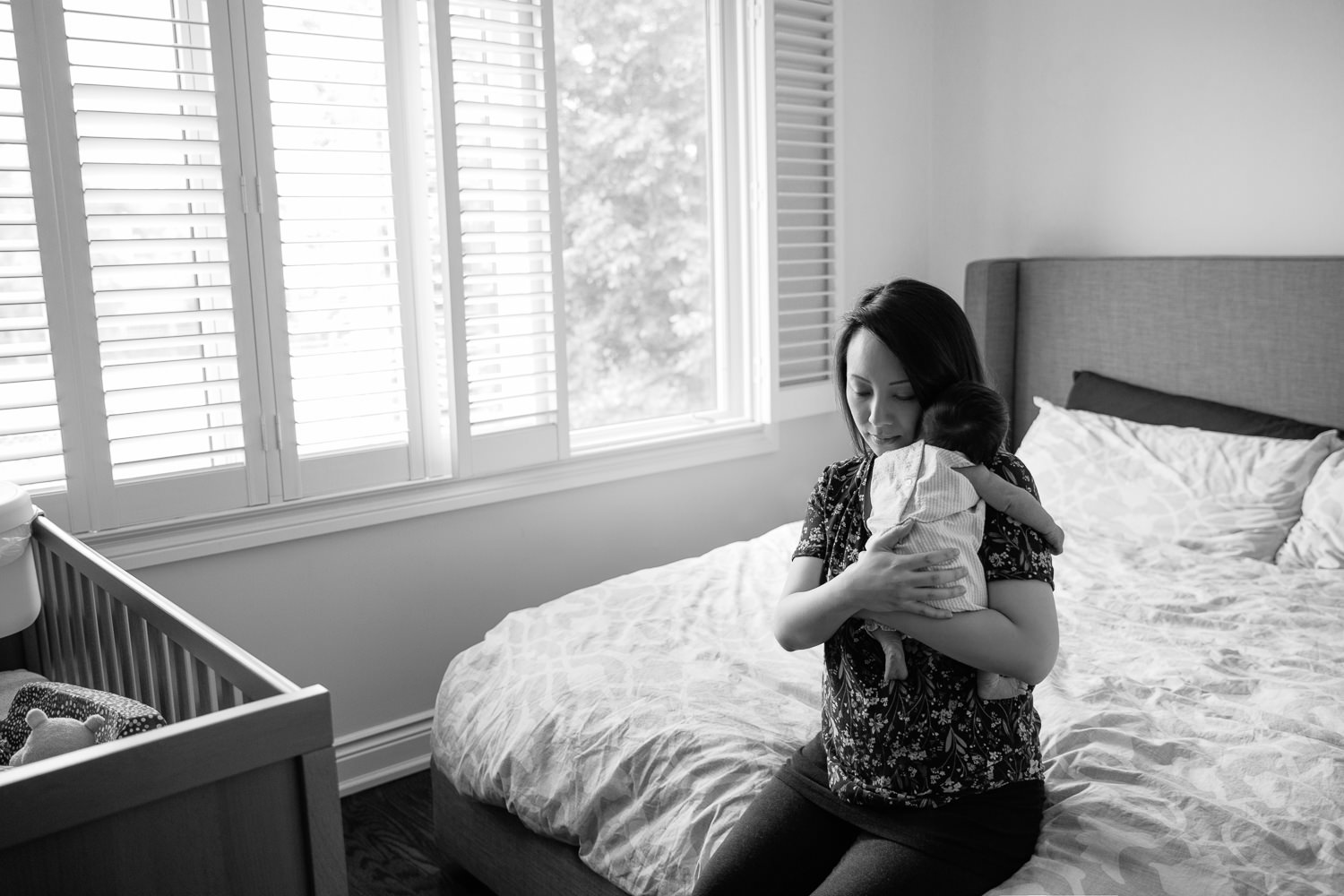 mom sitting on edge of bed snuggling sleeping 2 week old baby girl in white and purple striped outfit to her chest - Markham Lifestyle Photos