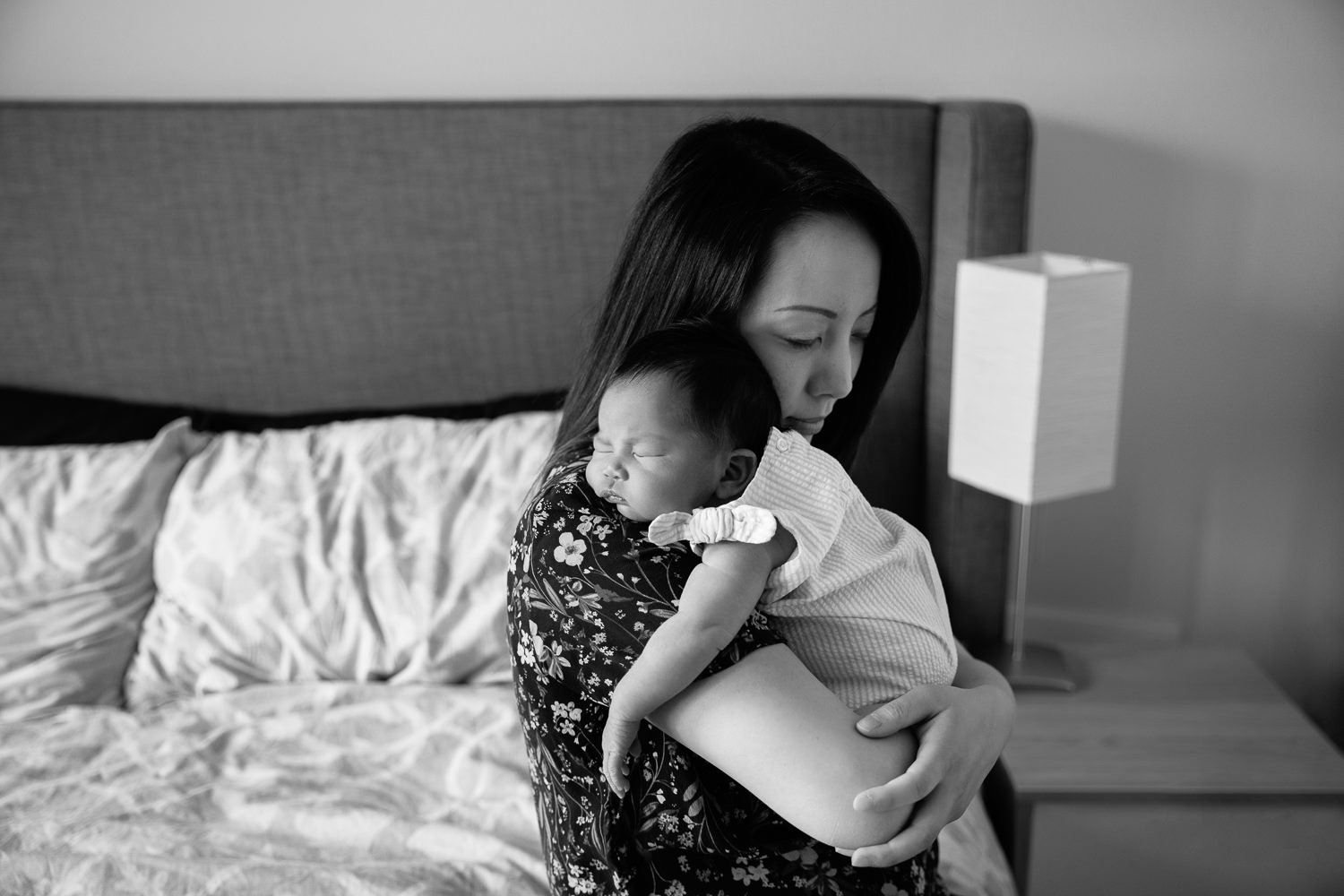 mom sitting on edge of bed snuggling sleeping 2 week old baby girl who has head resting on mother's shoulder - Barrie Lifestyle Photos