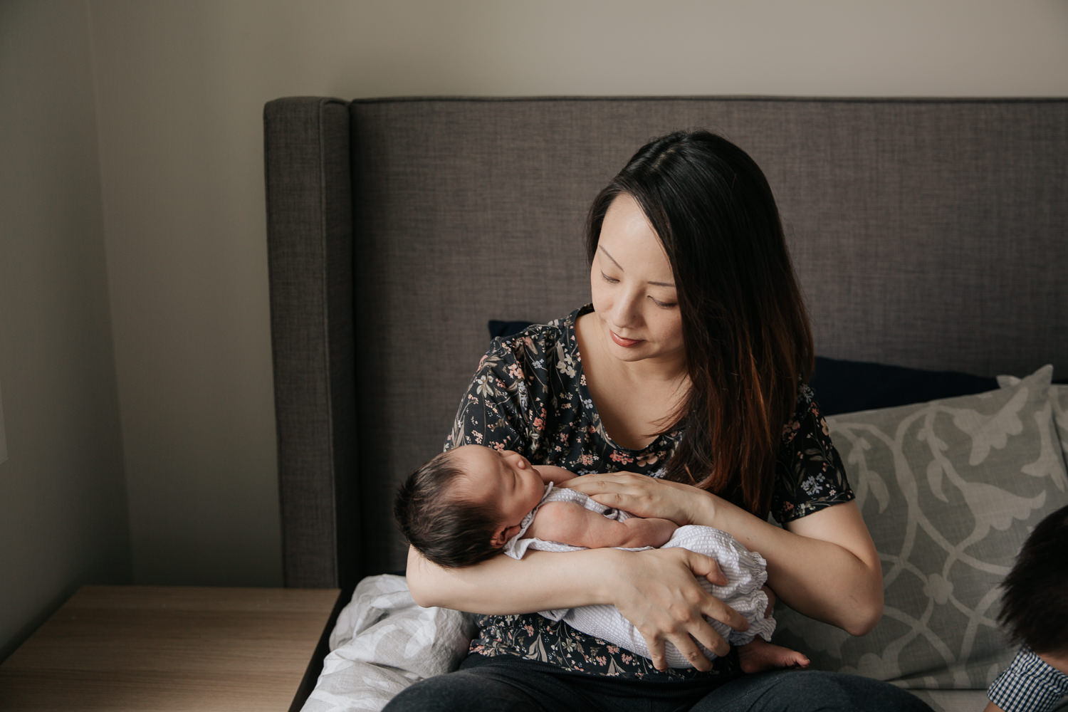 mom with long dark hair wearing floral top sitting on master bed holding and looking at 2 week old baby girl in her arms - Newmarket Lifestyle Photography