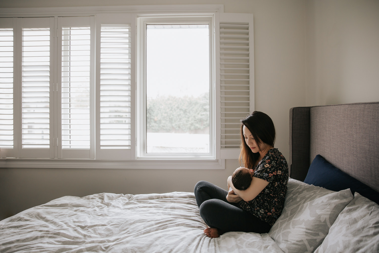 mom with long dark hair wearing floral top sitting cross-legged on master bed in front of window holding 2 week old baby girl in her arms - GTA Lifestyle Photos