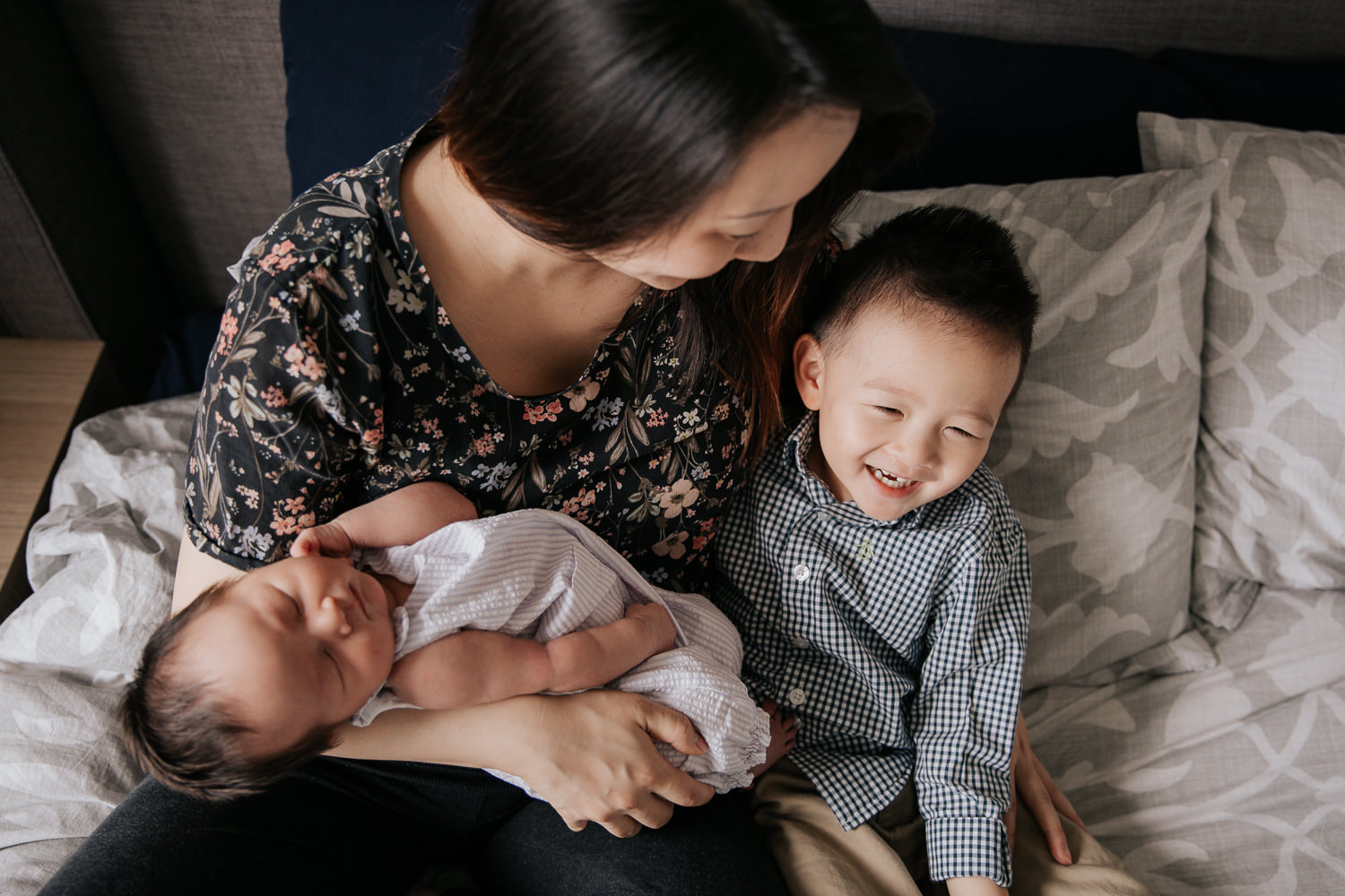 mom with long dark hair wearing floral top sitting on master bed holding 2 week old baby girl in her arms, snuggling and looking at smiling 3 year old toddler boy - Stouffville Lifestyle Photography