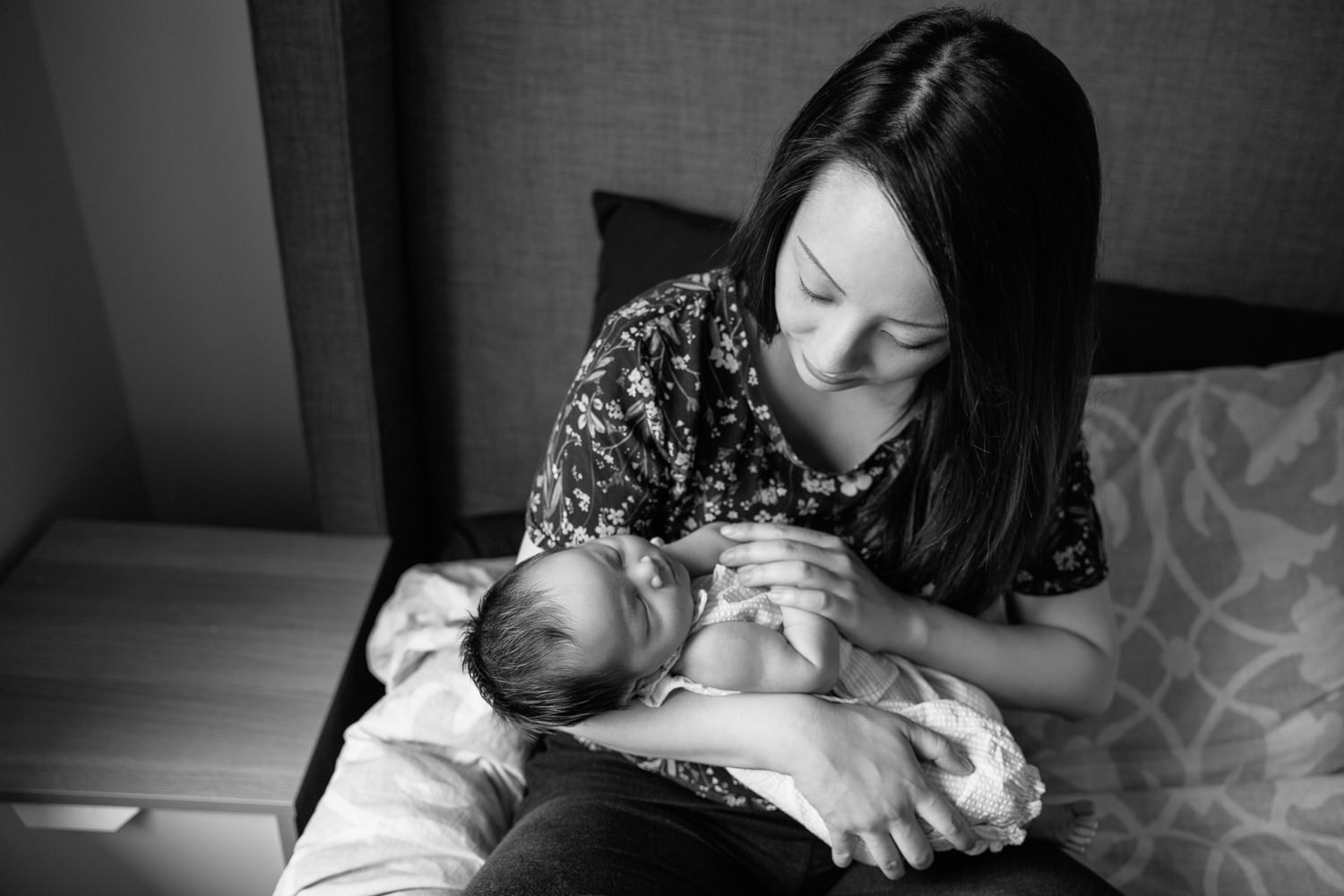 mom with long dark hair wearing floral top sitting on master bed holding and looking at 2 week old baby girl in her arms - Markham Lifestyle Photography