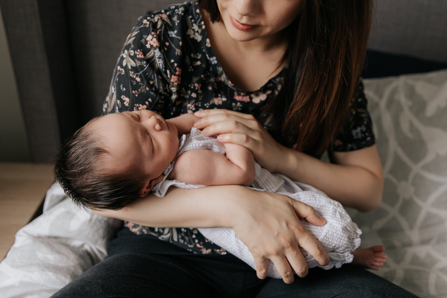 mom with long dark hair wearing floral top sitting on master bed holding and looking at 2 week old baby girl in her arms - Barrie Lifestyle Photography