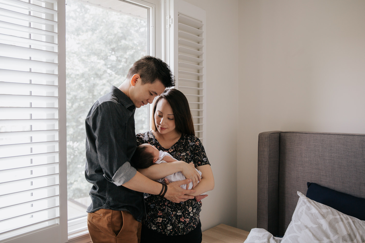 mom and dad standing in front of window holding sleeping 2 week old baby girl with dark hair in purple outfit, mother holding baby, father embracing them - Newmarket In-Home Photography