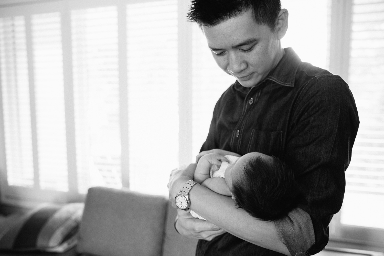 dad standing in front of window holding 2 week old baby girl in his arms, looking down at her - Stouffville In-Home Photography