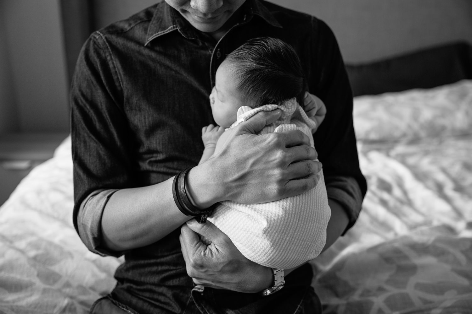 dad sitting on edge of bed holding sleeping 2 week old baby girl with dark hair to his chest, close up of hands holding baby - GTA In-Home Photography