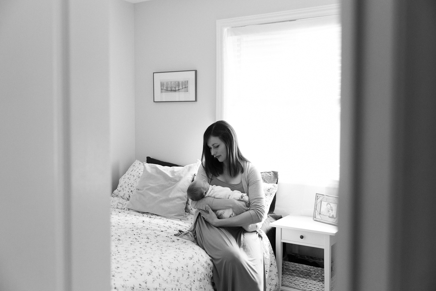 first time mother sitting on edge of master bed holding and looking at 3 week old swaddled baby boy in her arms - Stouffville Lifestyle Photography