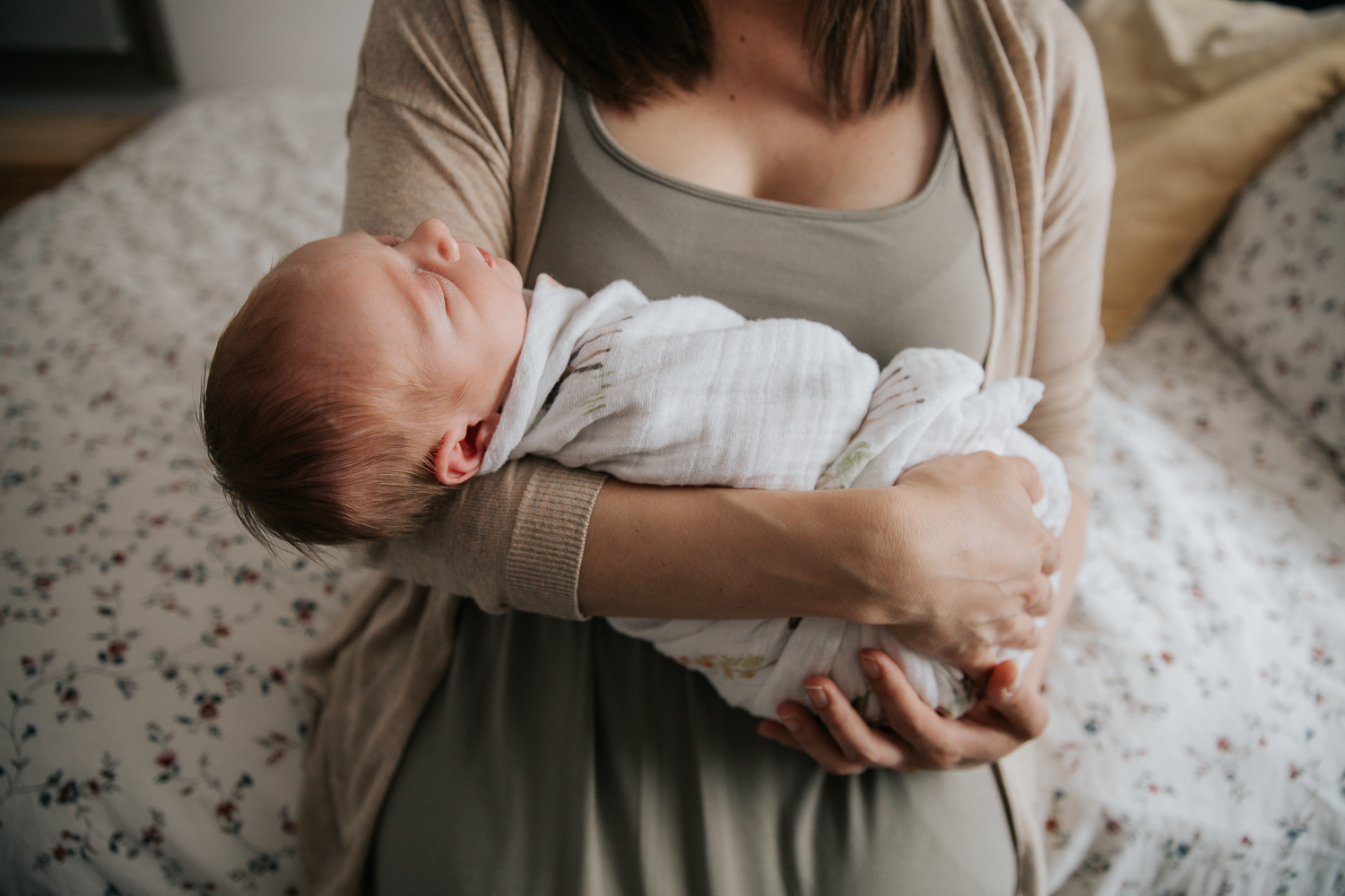 first time mother sitting on edge of master bed holding and looking at 3 week old swaddled baby boy with red hair in her arms - Markham Lifestyle Photography