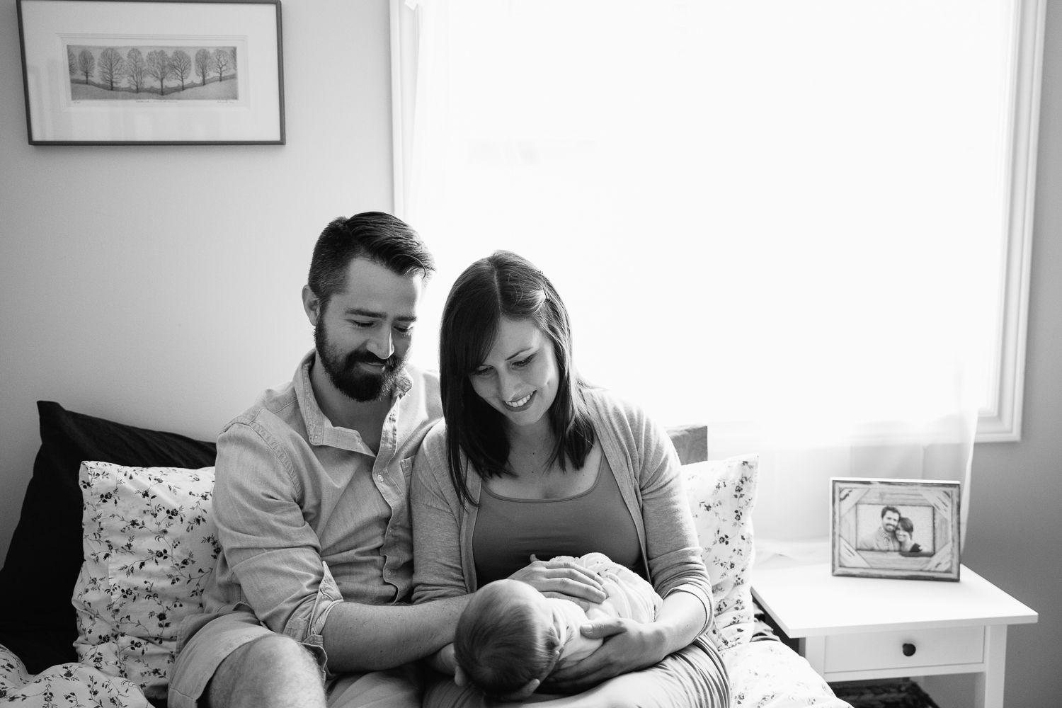 new parents sitting on master bed in front of window, mom holding 3 week old baby boy and dad with hand on his body, smiling at son -  GTA Lifestyle Photography