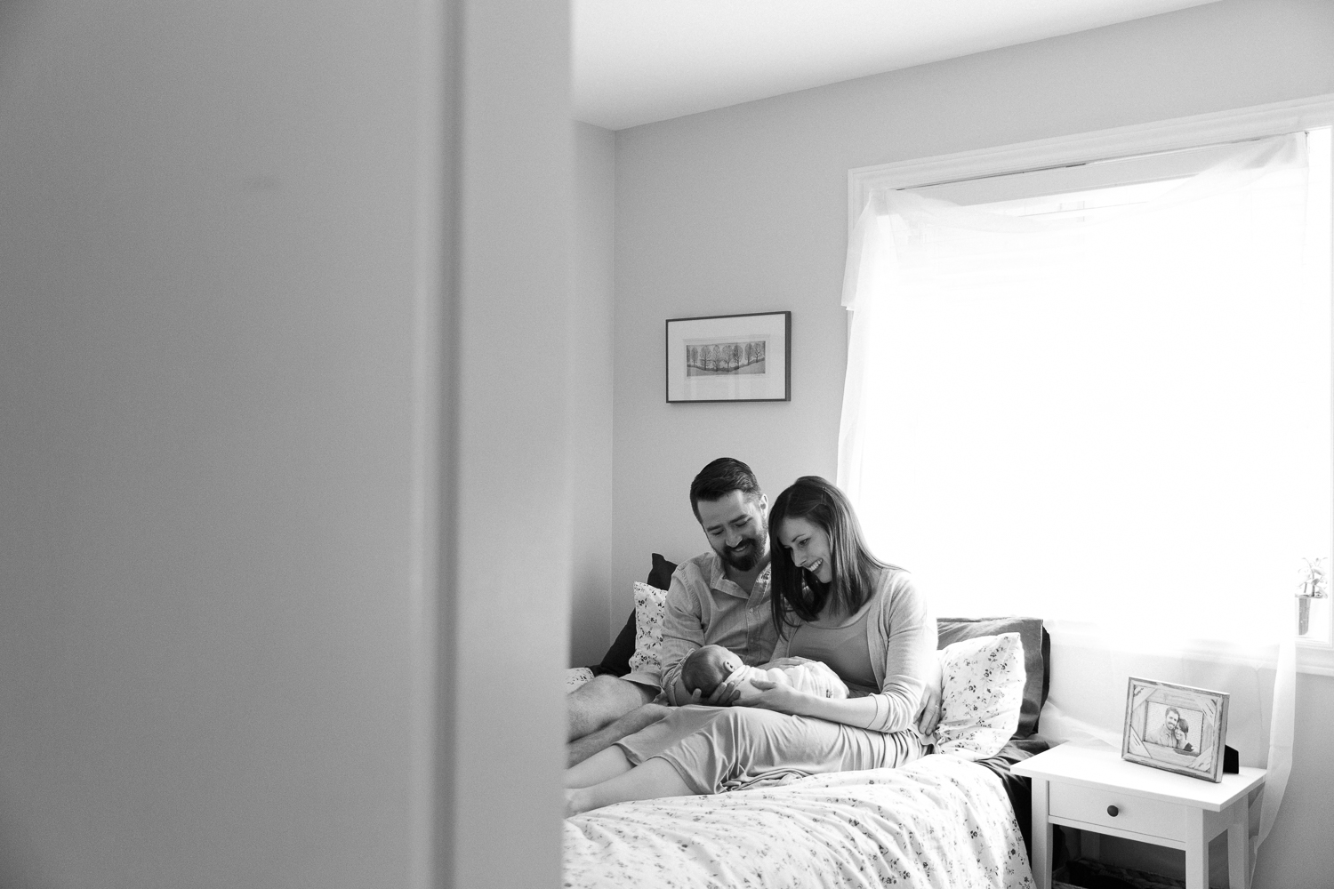 new parents sitting on master bed in front of window, mom holding 3 week old swaddled baby boy and dad with hand on his body, smiling at son - Stouffville In-Home Photography