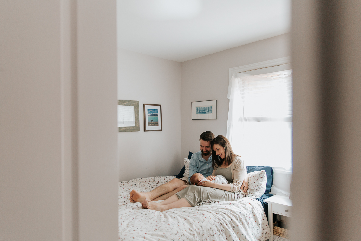 new parents sitting on master bed in front of window, mom holding 3 week old swaddled baby boy with red hair and dad with hand on his body, smiling at son - Newmarket In-Home Photography