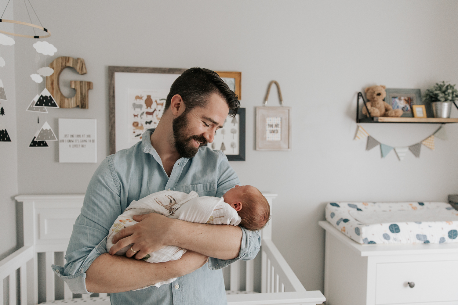 new father with brown hair and beard standing in nursery in front of crib holding 3 week old swaddled baby boy with red hair in his arms and smiling at son - Newmarket Lifestyle Photos