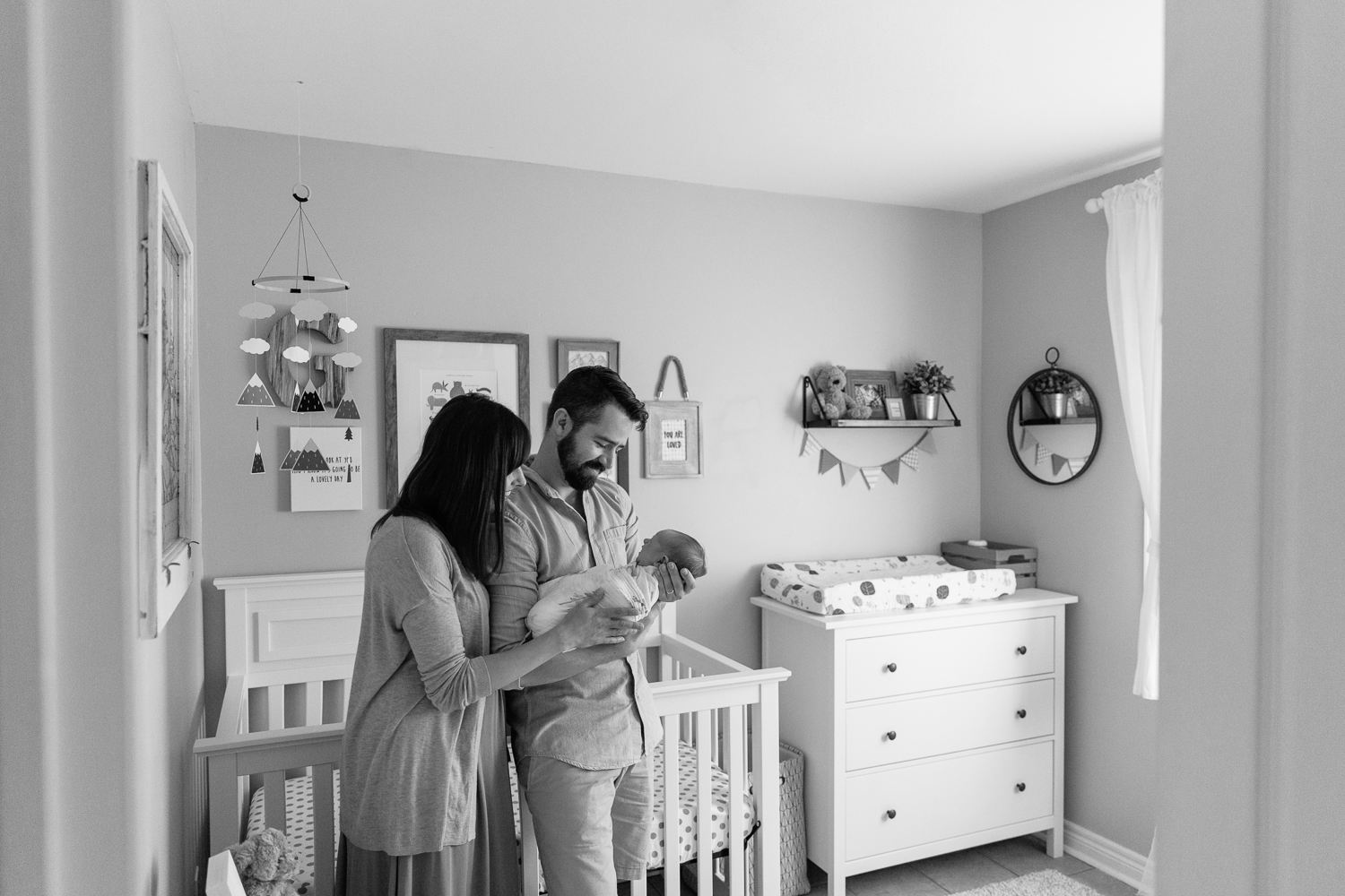 new parents standing in nursery in front of crib, dad holding and looking at swaddled 3 week old baby boy, mom standing next to husband with hand on son - Stouffville Lifestyle Photography