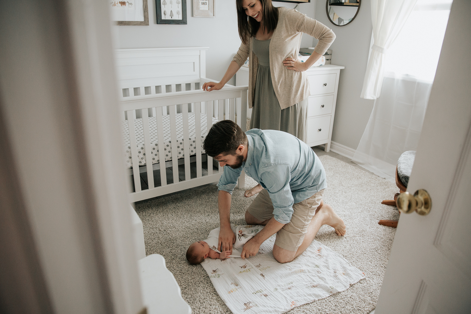 first time dad crouching on ground swaddling 3 week old baby boy on nursery floor while mom leans on crib watching and smiling - Newmarket Lifestyle Photography