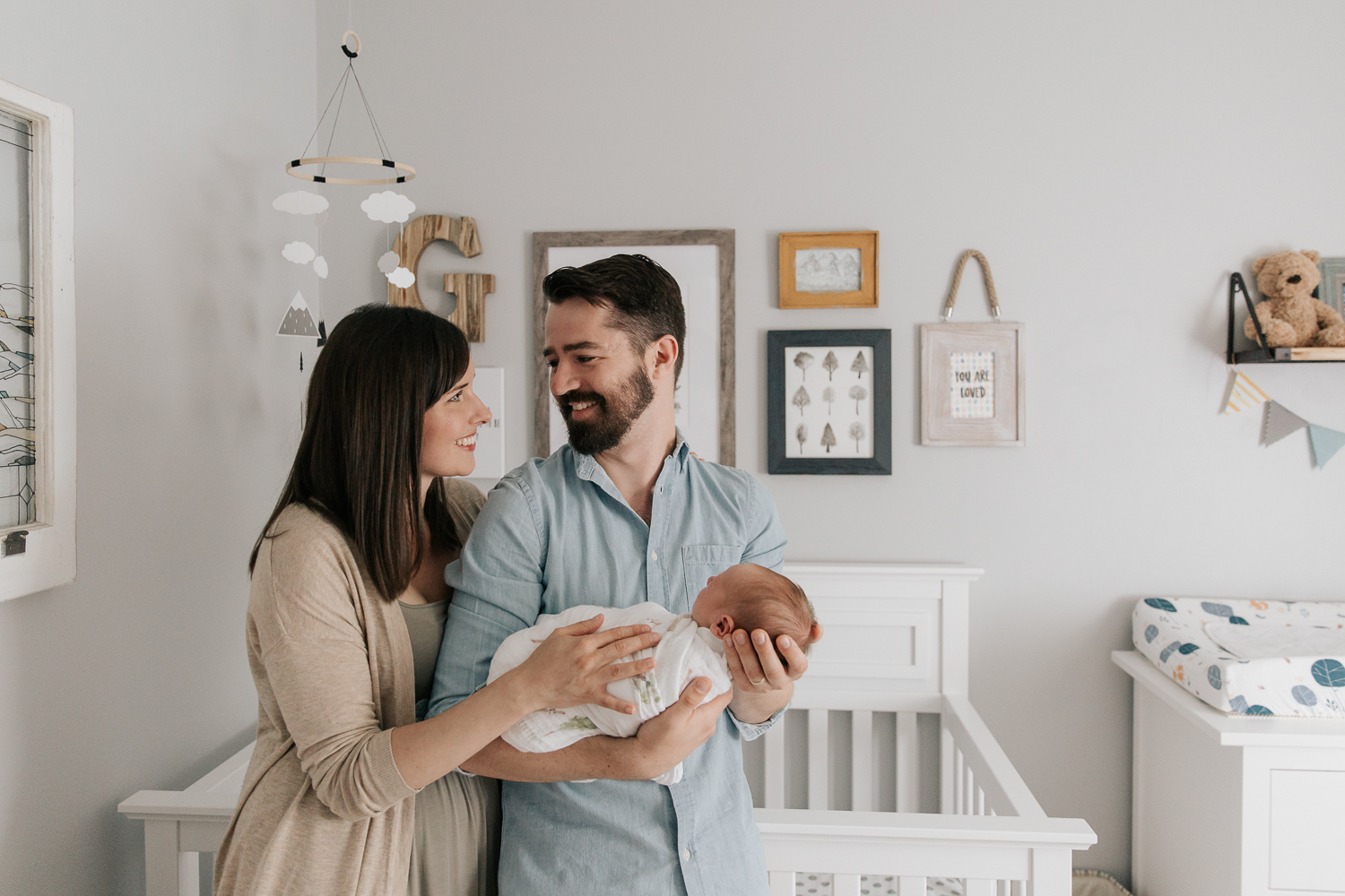 new parents standing in nursery in front of crib, smiling at one another, dad holding swaddled 3 week old baby boy, mom standing next to husband with hand on son - Markham Lifestyle Photography