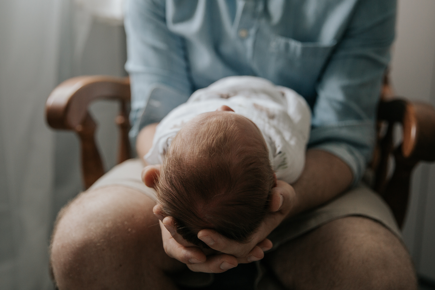 new father in blue button down shirt sitting in nursery glider holding sleeping 3 week old baby boy with red hair - Barrie In-Home Photography