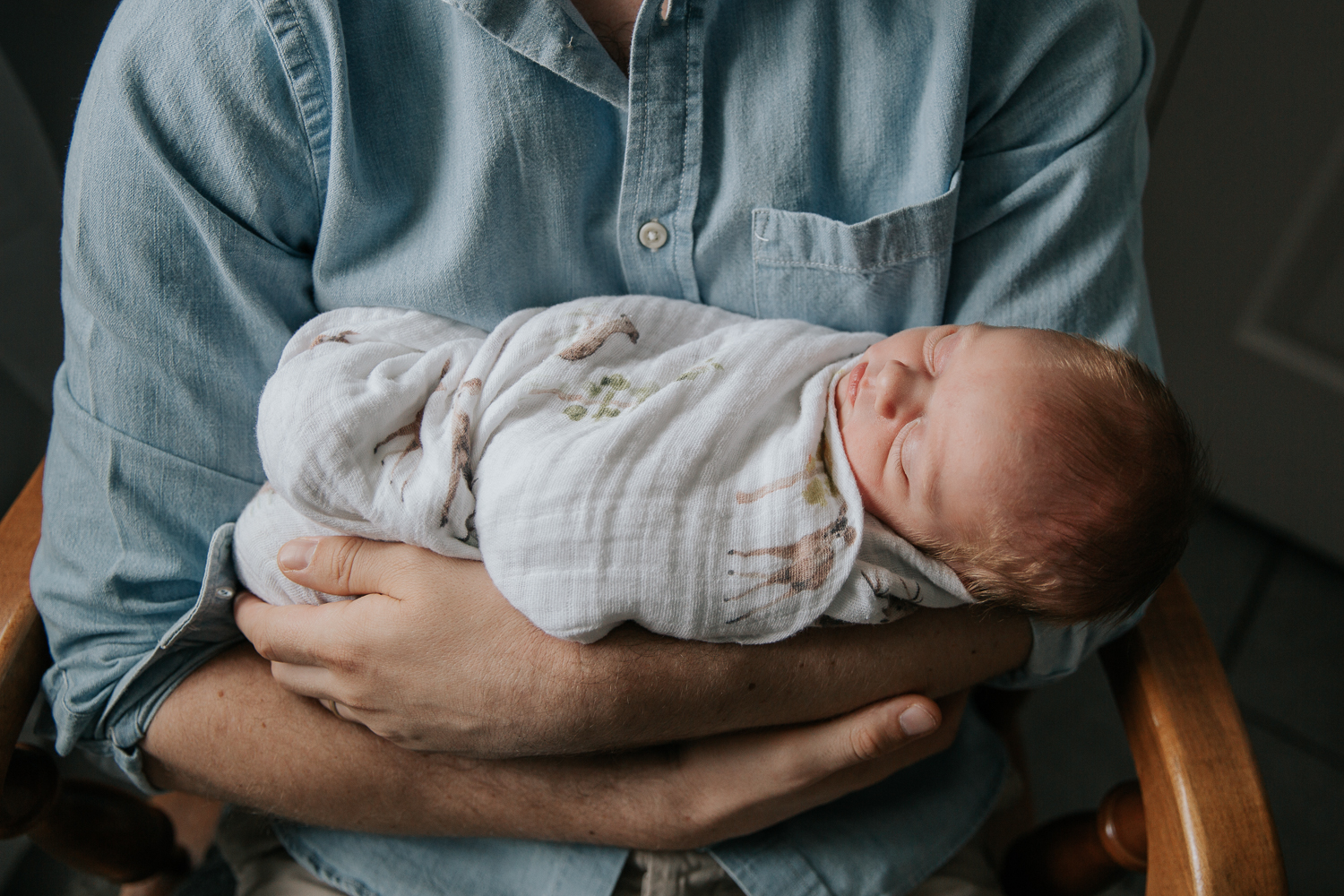 new father in blue button down shirt sitting in nursery glider holding sleeping 3 week old baby boy with red hair - Stouffville In-Home Photography