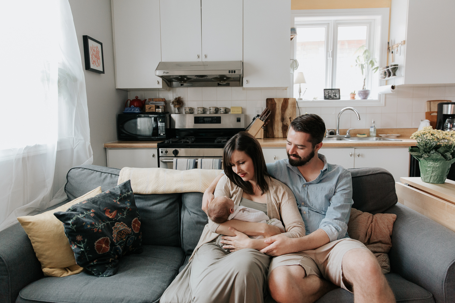 family of 3, new parents sitting on couch, mom holding 3 week old swaddled baby boy with red hair, dad with arm around mom looking at son - Newmarket Lifestyle Photos