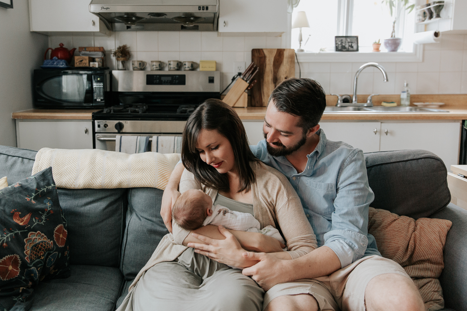 family of 3, new parents sitting on couch, mom holding 3 week old swaddled baby boy with red hair, dad with arm around mom looking at son - Stouffville Lifestyle Photos