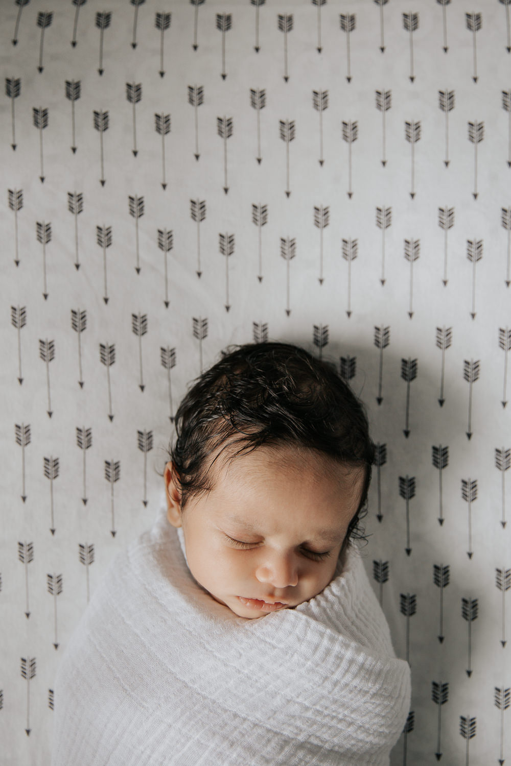 1 month old baby boy with dark hair wrapped in white swaddle lying asleep in crib on black and white arrow patterned sheet, head tilted to one side - Markham Lifestyle Photography