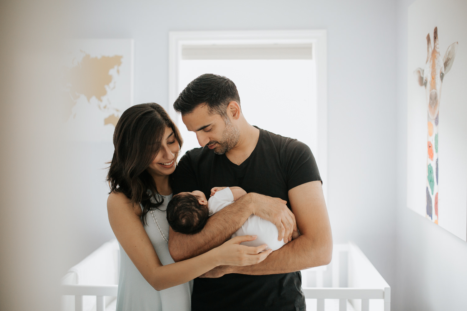 family of 3, new parents standing in nursery, dad holding 1 month old baby boy in onesie with dark hair, mom standing behind with arm wrapped around husband - Barrie In-Home Photography