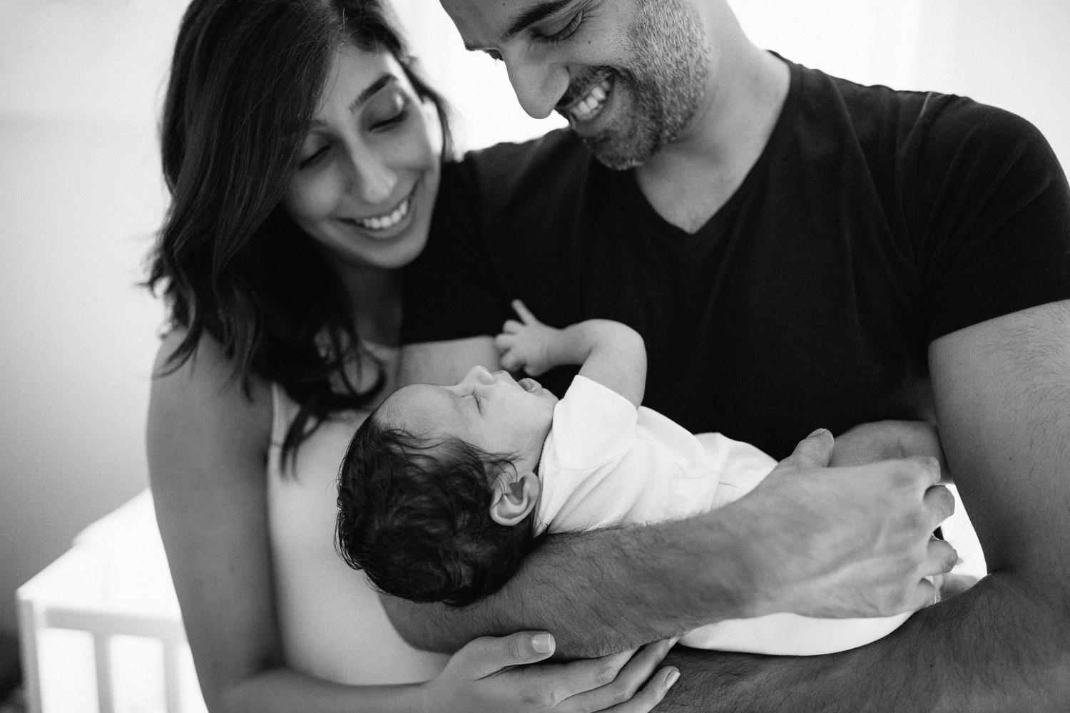 new parents standing in nursery, dad holding 1 month old baby boy in onesie with dark hair, mom standing behind with arm wrapped around husband, both smiling at son - Markham In-Home Photography