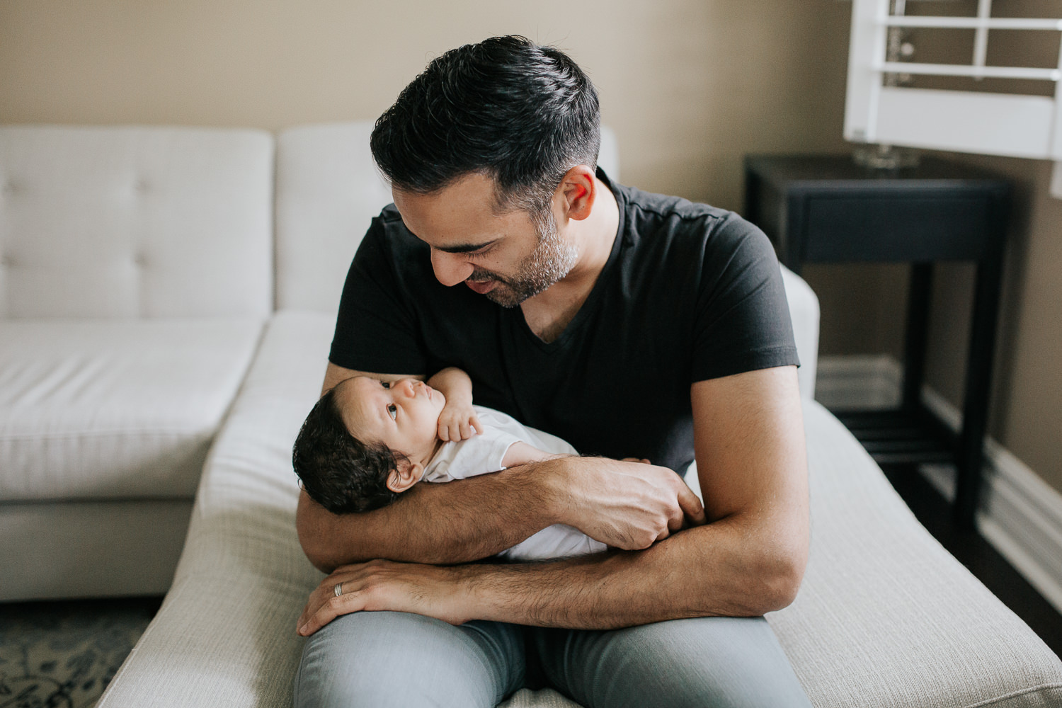 new father sitting on edge of couch holding 1 month old baby boy in onesie in his arms and talking to son - Newmarket In-Home Photography