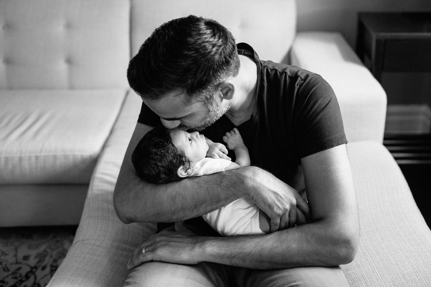 new father sitting on edge of couch holding 1 month old baby boy in onesie in his arms and kissing son on forhead - York Region Lifestyle Photography