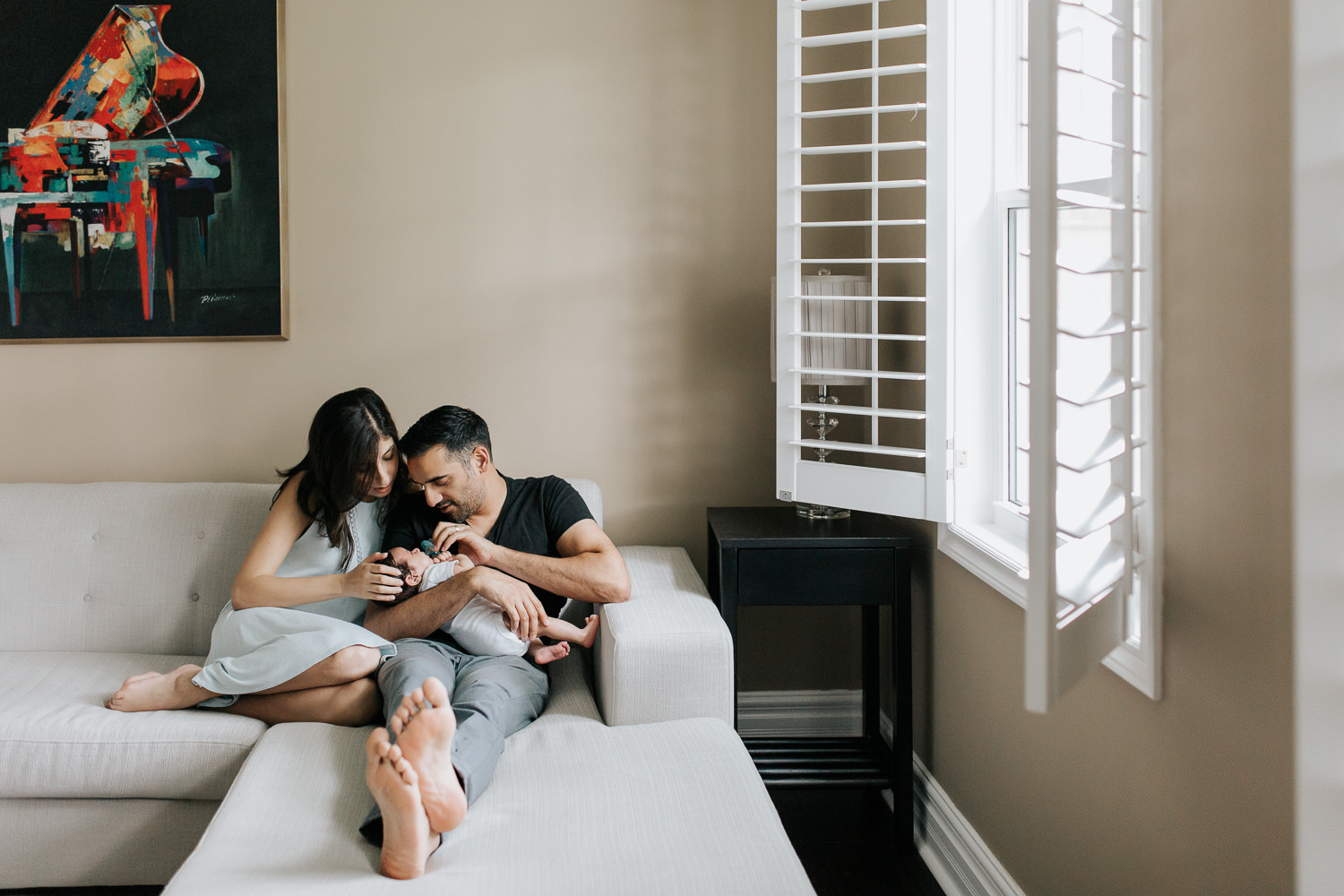 new parents sitting on sectional couch, dad holding 1 month old baby boy in onesie with dark hair, mom snuggled up next to husband with hand on son's head - Barrie In-Home Photos