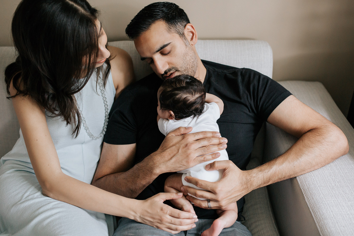 new parents sitting on couch, dad holding 1 month old baby boy in onesie with dark hair, mom snuggled up next to husband with hand on son's foot - GTA In-Home Photos