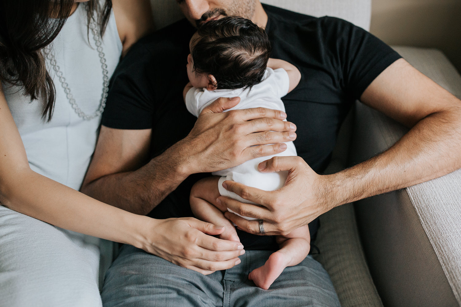 new parents sitting on couch, dad holding 1 month old baby boy in onesie with dark hair, mom snuggled up next to husband with hand on son's foot - York Region In-Home Photos