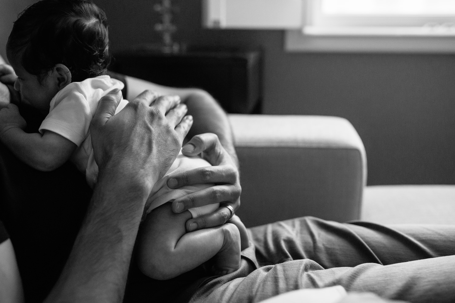 father sitting on couch with 1 month old baby boy in onesie lying on his chest, close up of hands on son - Newmarket Lifestyle Photos