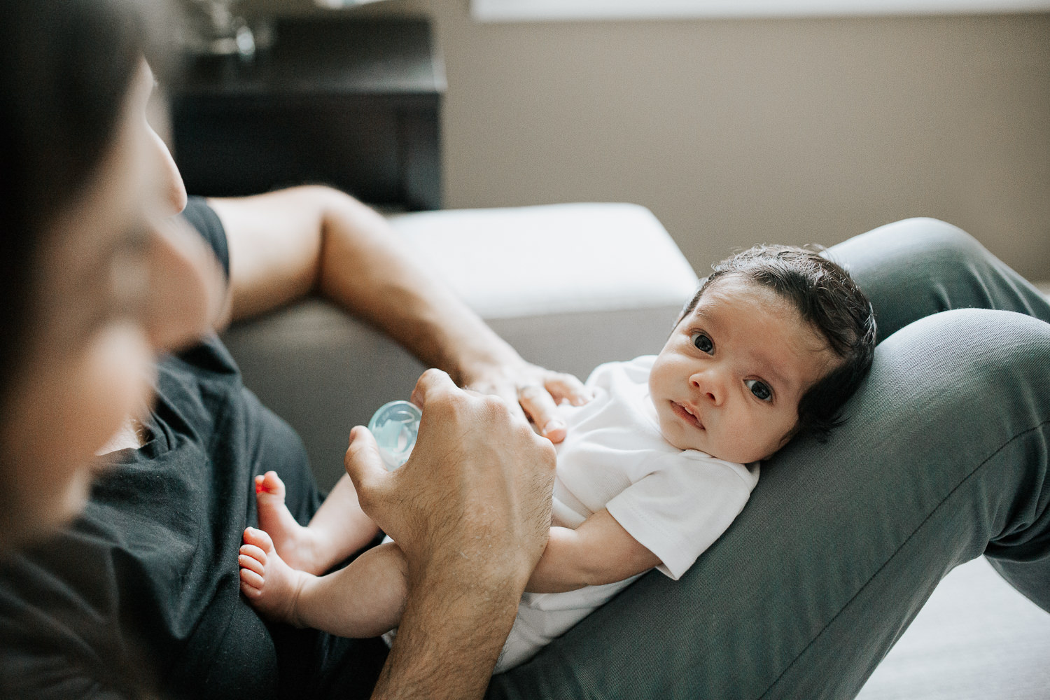 new parents sitting on couch, 1 month old baby boy in onesie with dark hair lying on dad's lap, baby looking at camera - Markham Lifestyle Photos