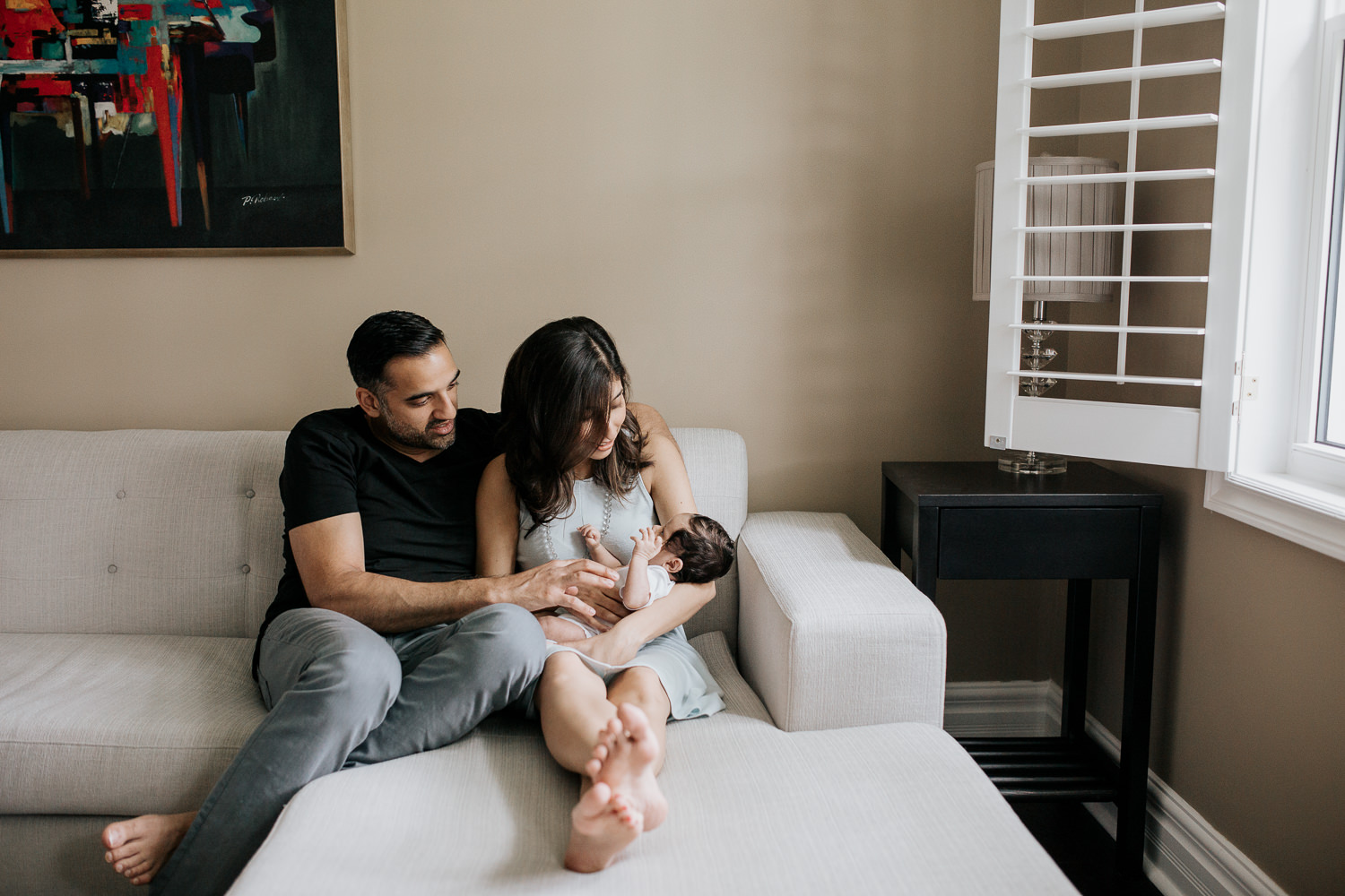 new mom sitting on couch snuggling 1 month old baby boy with dark hair in her arms, dad sitting let to wife with hand on son - Stouffville Lifestyle Photography