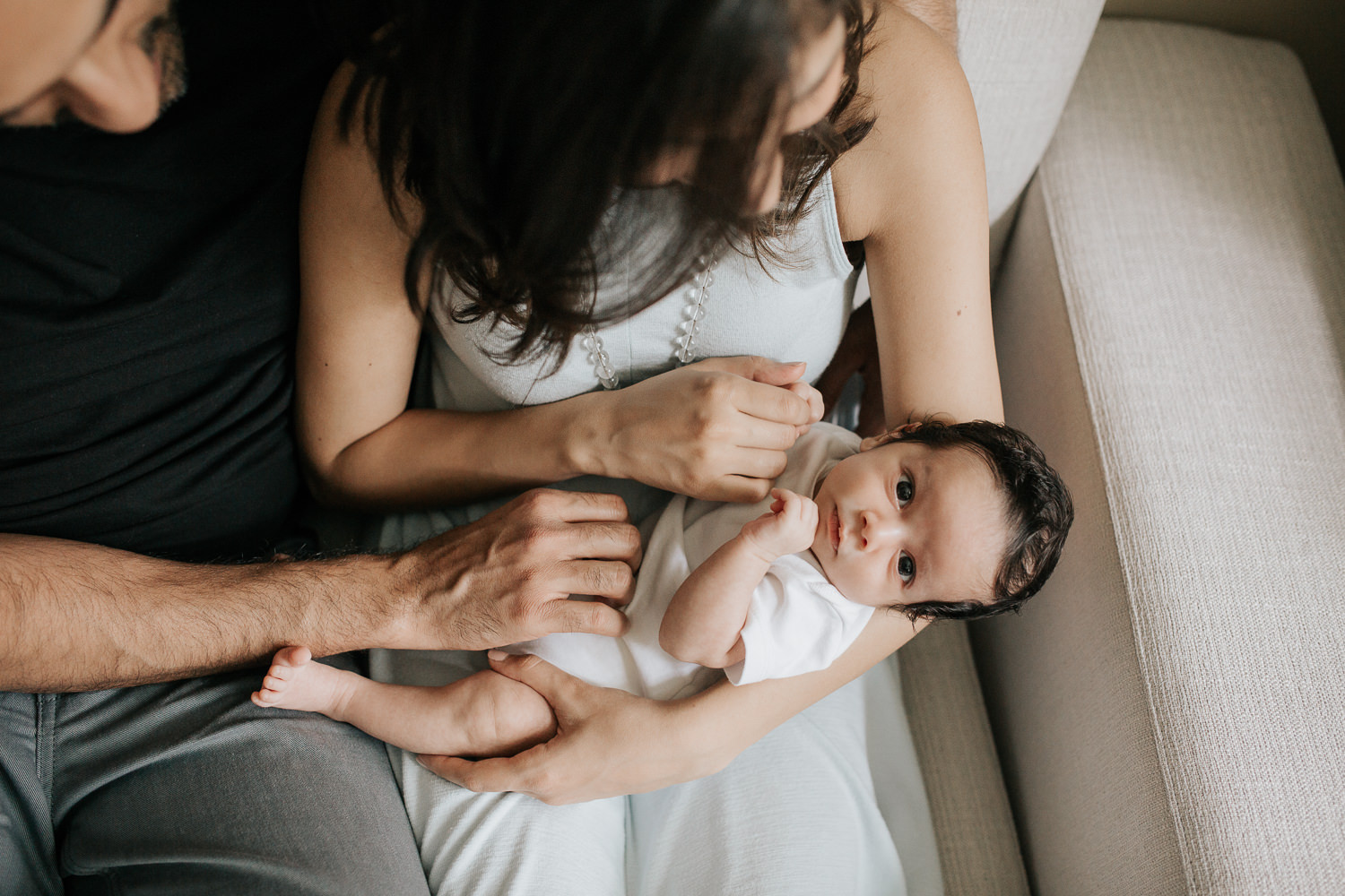1 month old baby boy in white onesie with lots of dark hair lying in mom's nap, looking at camera, mother holding son's hand and dad resting hand on baby - Markham Lifestyle Photography