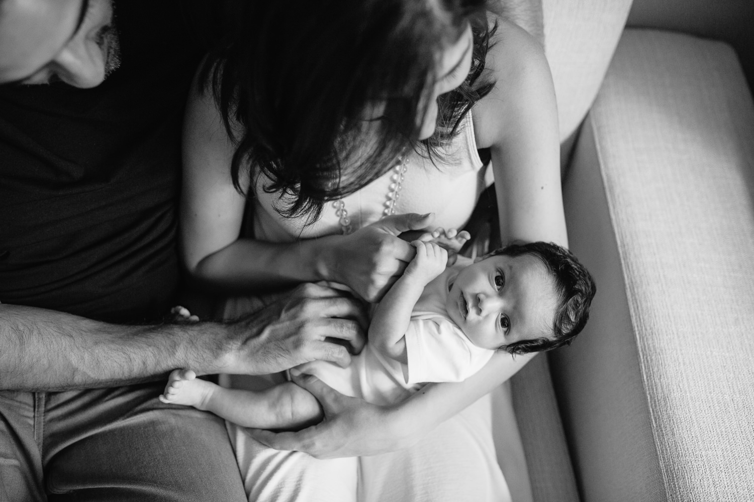 1 month old baby boy in white onesie with lots of dark hair lying in mom's nap, looking at camera, mother holding son's hand and dad resting hand on baby - Barrie Lifestyle Photography