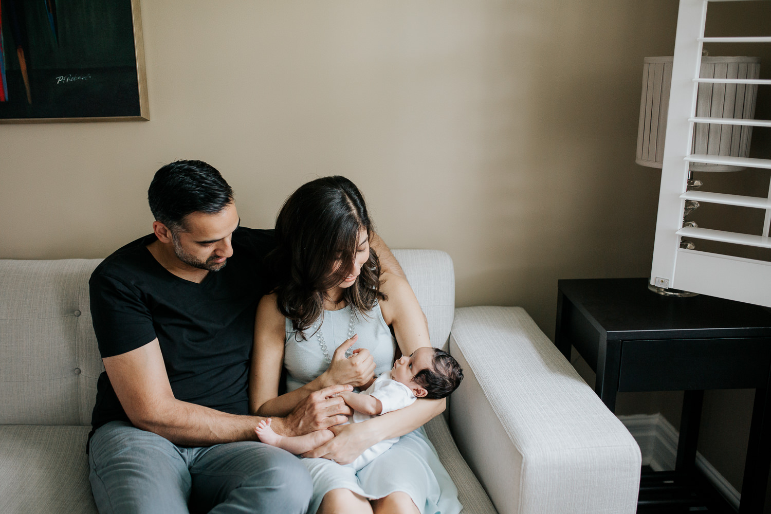 new mom sitting on couch snuggling 1 month old baby boy with dark hair in her arms, dad sitting let to wife with hand on son - GTA Lifestyle Photography