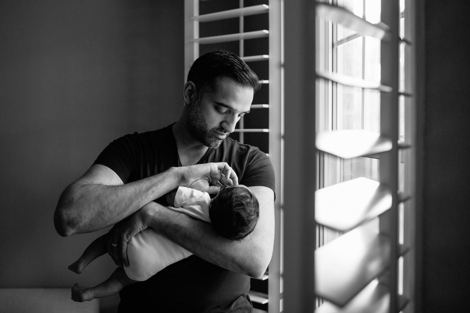 new father standing in front of window holding 1 month old baby boy in onesie in his arms, putting soother in son's mouth - York Region In-Home Photography