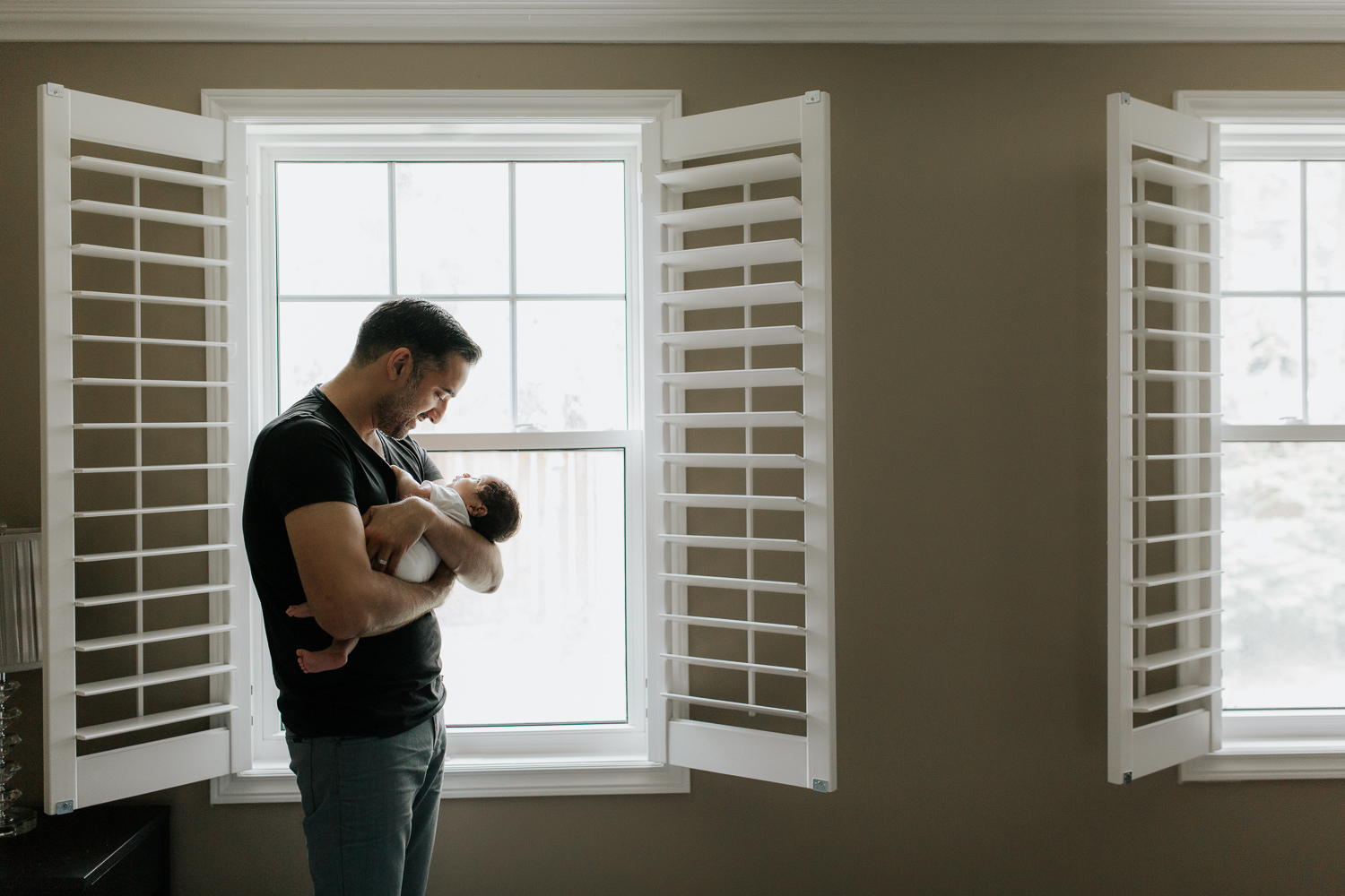 new father standing in front of window holding and smiling at 1 month old baby boy sleeping in his arms - GTA In-Home Photography