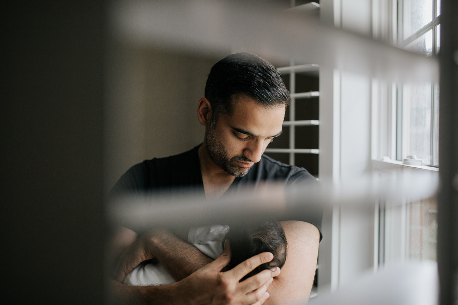 new father standing in front of window holding 1 month old baby boy in onesie in his arms, hand on son's head - Newmarket In-Home Photos